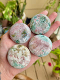Four polished Unicorn Jasper palm stones in pink, green, and creamy tones resting in a hand