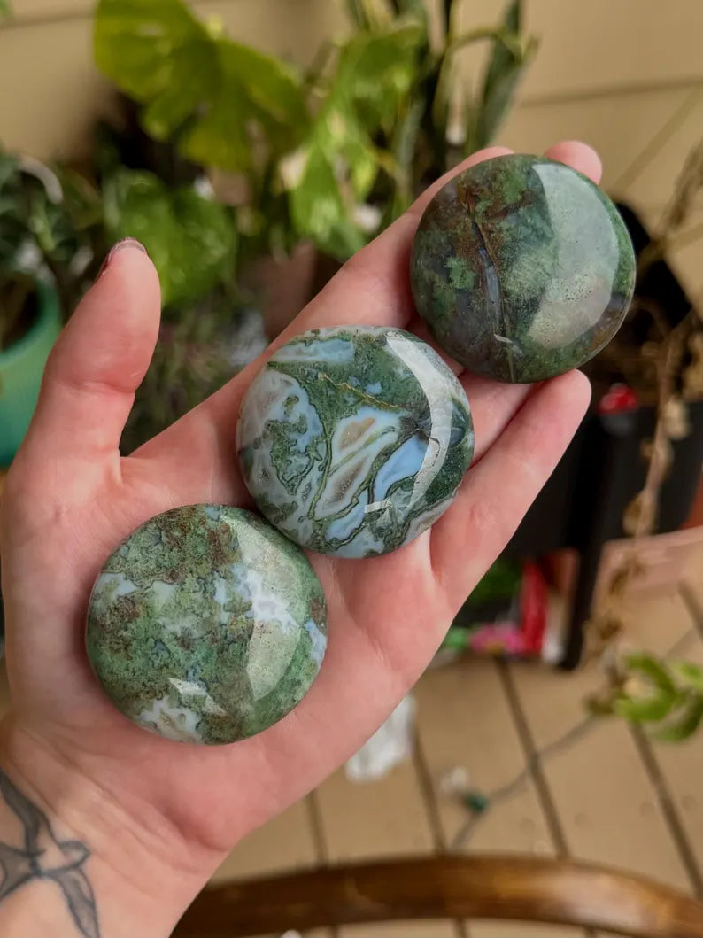 Three polished moss agate palm stones with green, gray, and creamy landscape-like patterns held in a hand